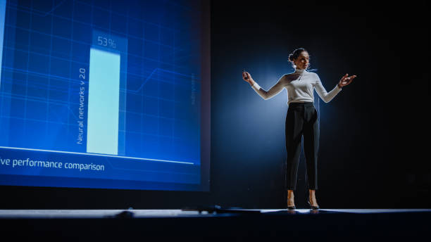 on-stage successful female speaker presents technological product, uses remote control for presentation, showing infographics, statistics animation on screen. live event / device release. - stage imagens e fotografias de stock