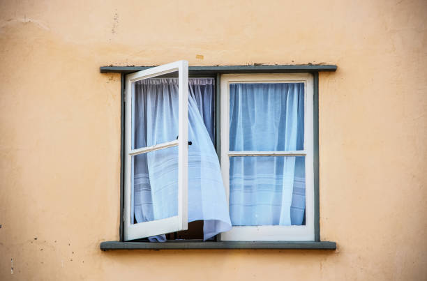 Open casement window in old stucco house with white gauze sheer curtains blowing out of them - close-up stock photo