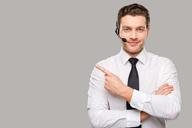 Operator pointing copy space. Handsome young man in formalwear and headset looking at camera and pointing away while standing against grey background call center photos stock pictures, royalty-free photos & images