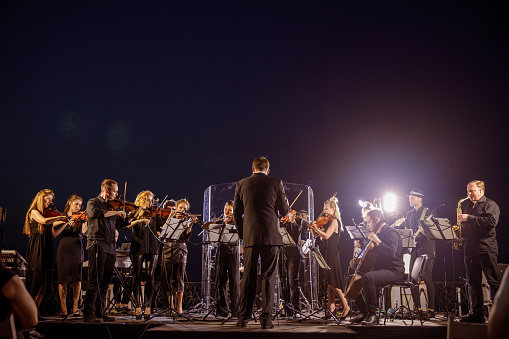 Musicians playing violin, violoncello, guitar and saxophone while conductor directing orchestra performance at night outdoor concert