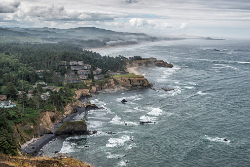 Overlook of the sandy beach and sea stacks along the Oregon Coast with grass bluff in the foreground and clouds and mountains in the background.
