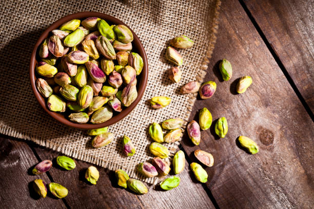 Organic pistachios still life Top view of a brown bowl filled with organic pistachios shot on rustic wood table. Some almonds are out of the bowl on a burlap. Predominant colors are green and brown. DSRL studio photo taken with Canon EOS 5D Mk II and Canon EF 100mm f/2.8L Macro IS USM pistachio stock pictures, royalty-free photos & images