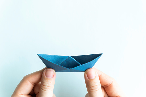 Hand is holding blue origami paper boat in front of white background.