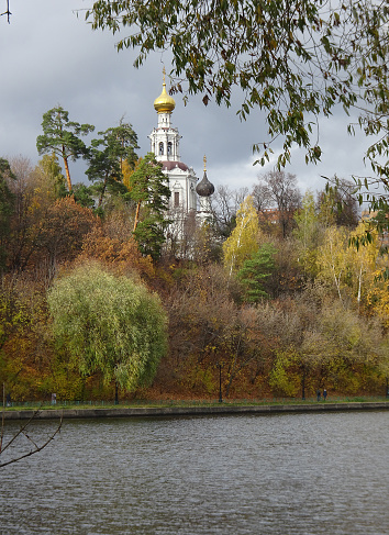 Colorful autumn. Alley along the riverbank on the background of the autumn park. Reflections in the water of the forest and blue sky. The domes of Orthodox churches dominate all