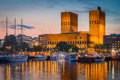 The iconic Functionalist towers of Oslo City Hall, Radhus, spotlit against the chrome blue dusk sky overlooking the harbour fjord and marinas of Aker Brygge, the popular leisure, dining and exclusive residential district in the heart of downtown Oslo, Norway's picturesque capital city. ProPhoto RGB profile for maximum color fidelity and gamut.