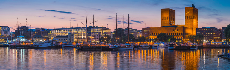 Summer sunset skies over the illuminated landmarks of Oslo's downtown waterfront, from the busy bars and restaurants of Aker Brygge, the popular leisure district, to the iconic twin towers of City Hall, Radhus, and the crowded marina on the tranquil waters of OsloFjorden, Norway.
