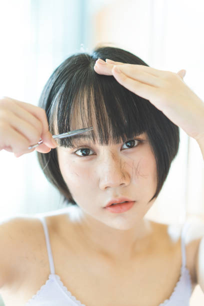 oung adult asian woman self cutting bangs haircut with scissors. Eyes looking at a camera stock photo