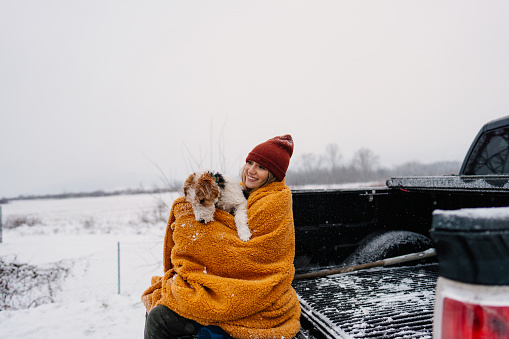 Photo of a young woman sitting in the back of a pickup truck with her puppy, wrapped in a blanket and taking a short break during their winter road trip.