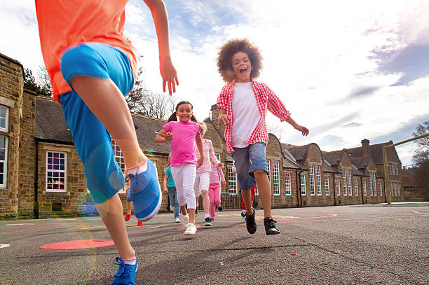 Children running across the school playground smiling. The children are running towards the camera. There are motion blurs and a sun flare. The children are casually dressed for summer. The school is visible in the background.
