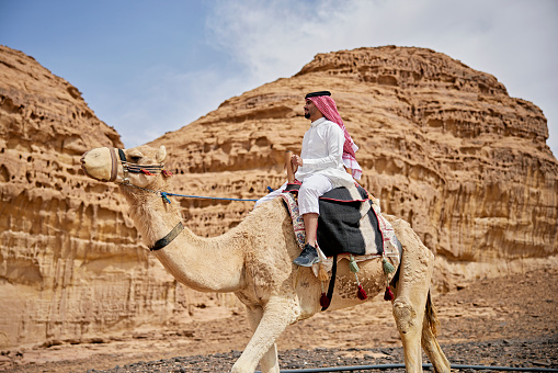 Side view of man in traditional Saudi dish dash, kaffiyeh, and agal sitting astride dromedary camel with red sandstone cliffs in background.