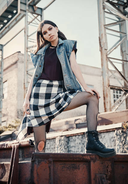 Outdoor portrait of pretty grunge (rock) girl at industrial place. Informal model dressed in jean jacket, checkered shirt, boots and holey tights stock photo