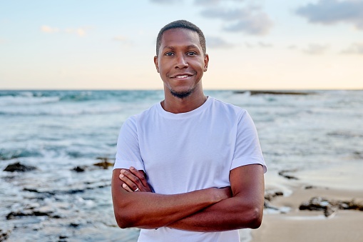 Outdoor portrait of a smiling young African American man looking into the camera. Positive confident 30s man with crossed arms, sky sea beach background. People, lifestyle, health, lifestyle concept
