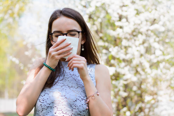 Outdoor shot of discontent young girl with seasonal allergy, uses white tissue, poses over blooming tree, has rhinitis and sneezing, reacts on allergens. People and disease concept Young girl has allergy for blooming spring trees antihistamine stock pictures, royalty-free photos & images