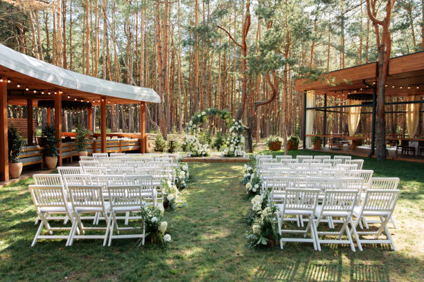 Outdoor wedding ceremony in the summer Outdoor Wedding Ceremony. A chic arch in the shape of a ring of white fresh flowers and green branches, white wooden chairs in a row on the grass. Pine forest in the background wedding ceremony stock pictures, royalty-free photos & images