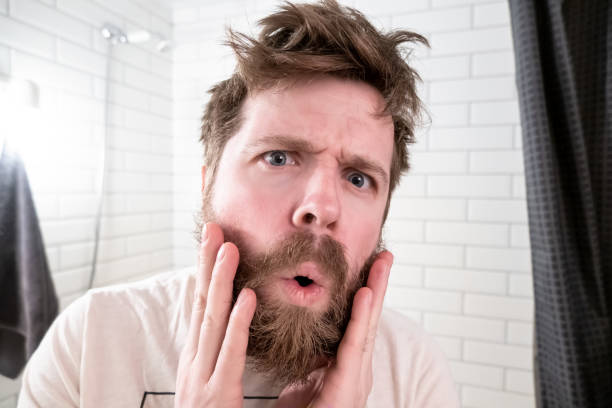 Overgrown man, with a shaggy hairstyle and beard, sees himself reflected in the mirror, he is shocked by his appearance. stock photo