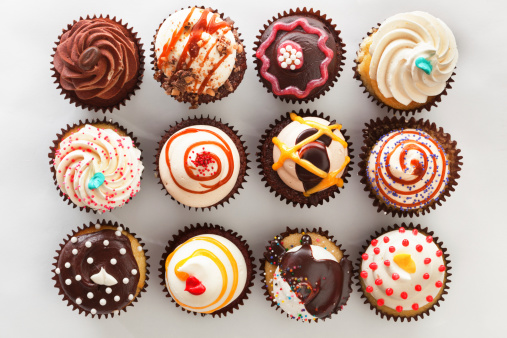 Subject: Horizontal high angle, directly overhead view of a dozen frosted cupcakes. The cupcakes are against a white background and are decorated with swirls, sprinkles, drizzles and candies—a tempting decadent treat for those who indulge in sweets and love cakes with lots of toppings.
