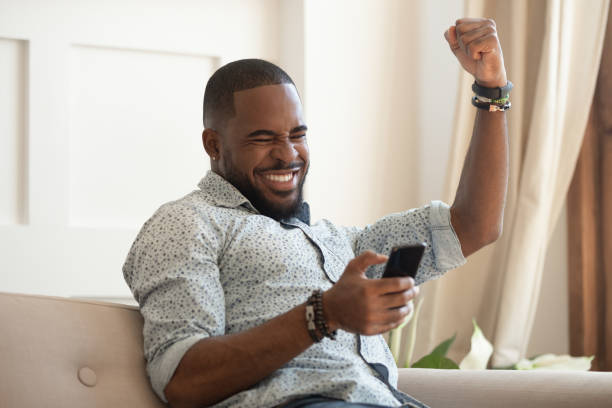 Overjoyed black man holding smartphone feeling euphoric with mobile win Excited overjoyed black man winner holding smartphone feeling euphoric with mobile online bet bid game win, happy ecstatic african guy looking at cell phone celebrate receiving reading good news incentive stock pictures, royalty-free photos & images