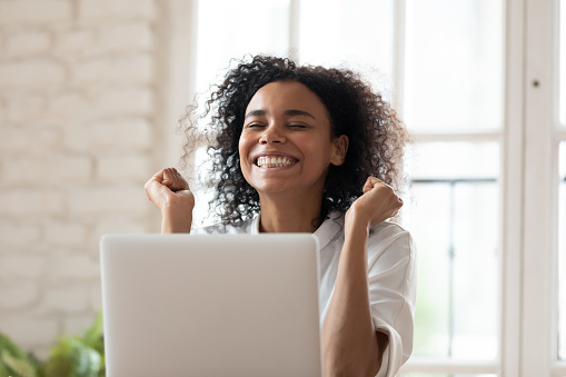 Overjoyed smiling young african ethnicity female employee feeling excited of reading email on computer with amazing news, celebrating success or achievement, job promotion, career opportunity concept.