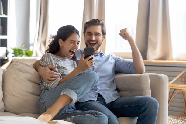 Overjoyed surprised young couple looking at phone screen Overjoyed surprised young couple looking at phone screen, reading message with good news, excited woman and man celebrating success, online lottery win, showing yes gesture, sitting on couch two parents stock pictures, royalty-free photos & images