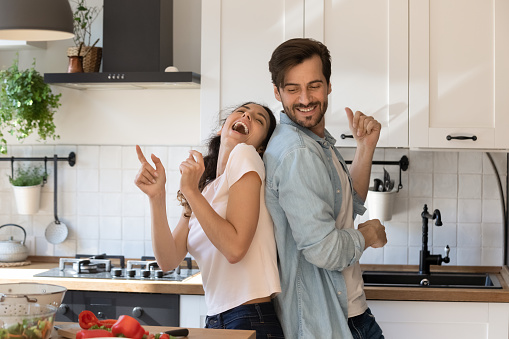Overjoyed young family having fun in modern kitchen, dancing and laughing, happy young wife and husband moving to favorite music, enjoying leisure time, cooking dinner together, romantic date