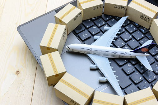 Overseas freight / international delivery or global logistics concept : White model airplane (heading left) lands on a laptop computer keyboard, encircled with light brown paper boxes or goods cartons