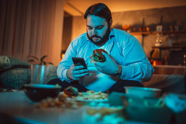 Overweight guy using phone One man, sitting at home, using mobile phone while watching movie. fat man looks at the phone stock pictures, royalty-free photos & images