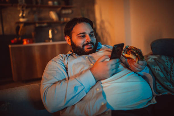 Overweight man looking at the phone One man, sitting at home, using mobile phone while watching movie. fat man looks at the phone stock pictures, royalty-free photos & images