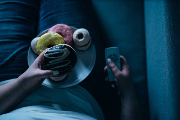 Mujer con sobrepeso tumbada en el sofá comiendo alimentos azucarados - foto de stock