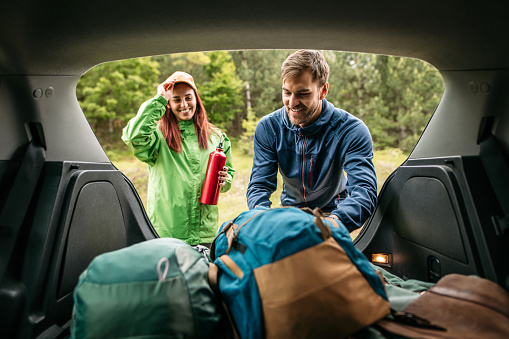 Photo of a smiling young couple, packing up camping equipment in the trunk of a car, ready for walking.