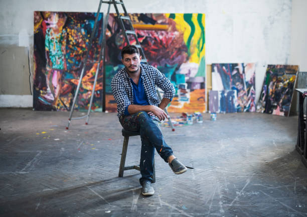 Painter Artist portrait in studio. Portrait of young artist painter sitting in front on his big modern paintings in art studio. artist stock pictures, royalty-free photos & images