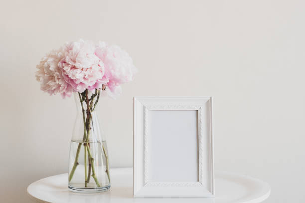 Pale pink peonies with blank rectangular picture frame on table Close up of pale pink peonies in glass vase with blank rectangular picture frame on white table - matte filter effect and selective focus vase photos stock pictures, royalty-free photos & images