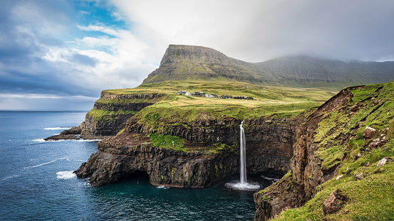Faroe Islands Panorama of the iconic natural Mulafossur - Múlafossur Waterfall and Gasadalur village on Vágar Island under blue summer sky with dark clouds over the coastal mountain range. Múlafossur Waterfall, Gásadalur - Gasadalur, Mykinesfjørður, Vagar Island, Faroe Islands, Denmark, Nordic Countries, Europe