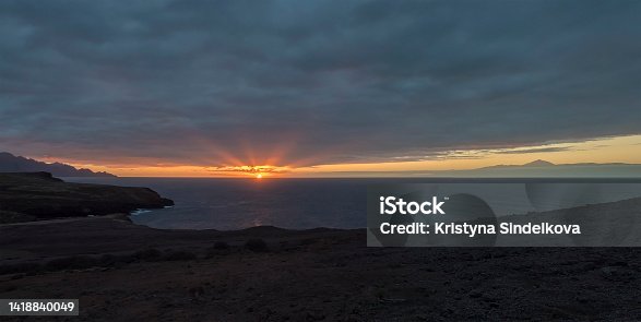 istock Panorama of beautiful sunset on the rocky atlantic coast in north west part of Gran Canaria island. Sun with rays going down to the ocean. Cliffs and Pico del Teide volacano in background. 1418840049