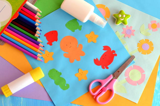 Paper marine animals and flowers cards. Sheets of colored paper, scissors, pensils, glue stick on a wooden background. Summer paper crafts for kids to make at home or in kindergarten stock photo