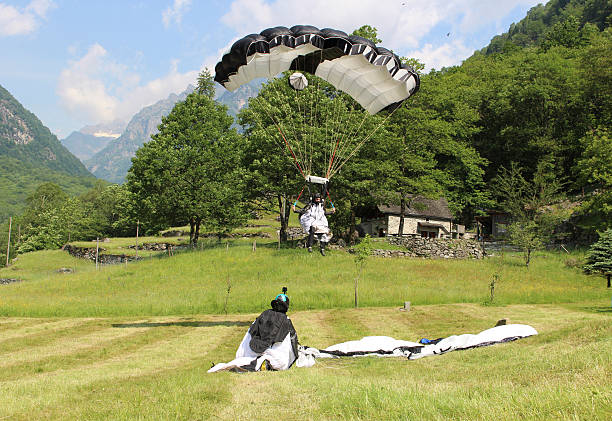 Parachuter comes in for touch down in Bern Canton, Switzerland. Parachuter comes in for touch down in Bern Canton, Switzerland. Mountains, forest and blue sky behind, while friend waits on field. base jumping stock pictures, royalty-free photos & images