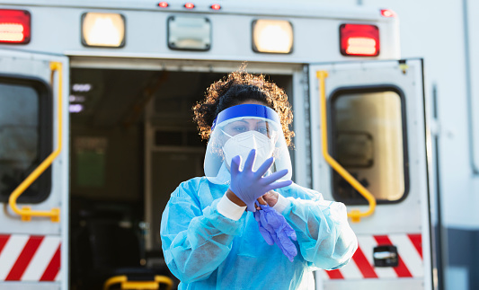 A paramedic standing in front of an ambulance, putting on PPE, or personal protective equipment, including a face mask, face shield, surgical gown and gloves. She is working during the COVID-19 pandemic, trying to protect herself from catching and spreading coronavirus. She is a mid adult woman in her 30s, mixed race Hispanic and Pacific Islander.
