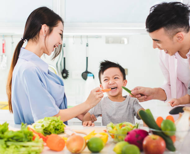 parent feeding boy a piece of  carrot in kitchen parent feeding boy a piece of  carrot in kitchen asian family eating together stock pictures, royalty-free photos & images