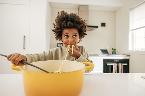 Cute child with afro eating spaghetti out of a big yellow pot in the kitchen. Child eating food with his hands.