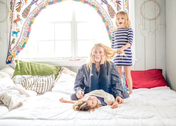 Parenting Mother in pajamas with coffee sits on the bed while children jump all around her bizarre stock pictures, royalty-free photos & images