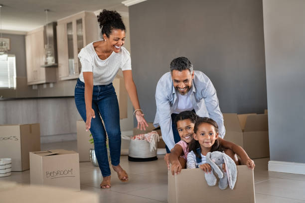 Parents playing with children while moving into new home Happy indian father playing with daughters sitting in carton box at new home. Happy multiethnic family having fun together in new house. Smiling dad pushing excited little girls in cardboard box after relocation. first time home buyer stock pictures, royalty-free photos & images