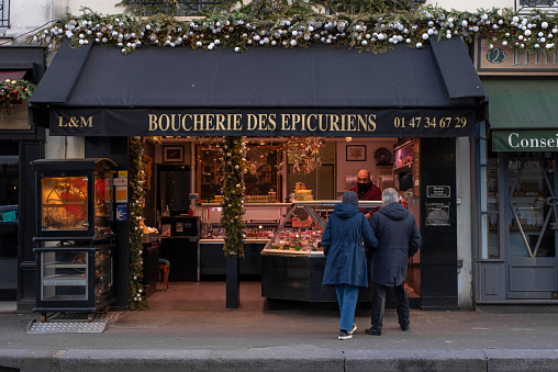 Paris, France - January 14 2022: Old couple standing in front of butchery shop and choosing fresh meat in Paris city centre on a cloudy winter morning.