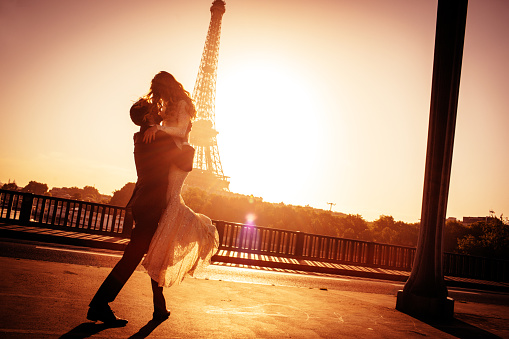 Young married couple is kissing on Pont d'Lena while sun is rising. There is an Eiffel tower in the background. 