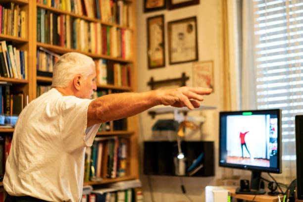 Parkinson Disease patient, standing in front of monitor, at home and online exercising dancing with help of a physical therapist Side view of Senior man, a Parkinson Disease patient, who is standing in front of monitor, at home, online exercising dancing with help of a physical therapist for Parkinson Disease. Online is only possible therapy as it is Covid-19 lockdown time. medical assistant online courses stock pictures, royalty-free photos & images