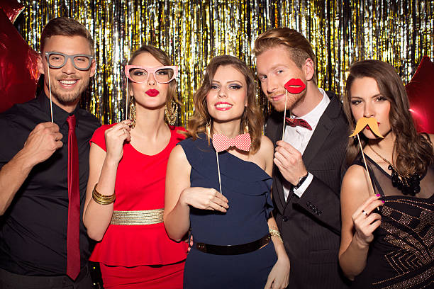 Party. Young people wearing elegant clothes celebrating or having party in front of fringe curtain. They are holding photo booth fake props as moustaches, glasses and kiss lips and posing at camera. formalwear photos stock pictures, royalty-free photos & images