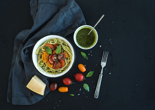 Pasta spaghetti with pesto sauce, basil, slow-roasted cherry-tomatoes in rustic metal bowl on dark grunge backdrop, top view, copy space