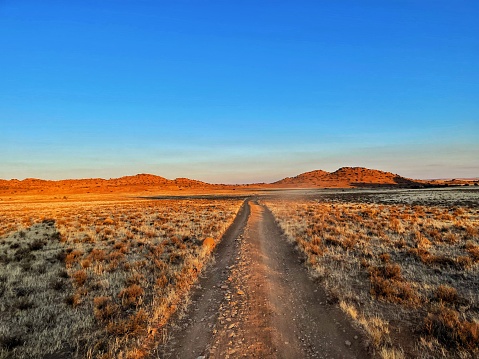 A semi-arid region, far from civilization, with plains stretching to the mountains on the horizon, with clouds covering the blue skies. A two-lane gravel road is showing the way to the horizon.