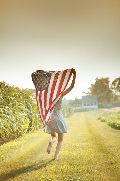 Patriotic Girl Flying American Flag, USA Fourth of July Banner Young farm girl running next to corn crop farm field, holding the USA American flag over her head, flying it behind her. The national banner is displayed for the Fourth of July, Memorial Day, and other political or patriotic holidays and events. Summer light glows on this rural Midwest landscape. memorial day stock pictures, royalty-free photos & images