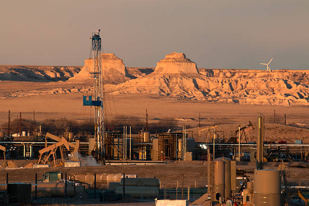 In the early morning along county road 127 on the plains in northern Colorado in Weld County, an oil drill rig / workover rig and multiple oil pump jacks, storage tanks and petroleum production equipment stand with the Pawnee National Grasslands, Pawnee Buttes and a wind turbine spinning in the background.