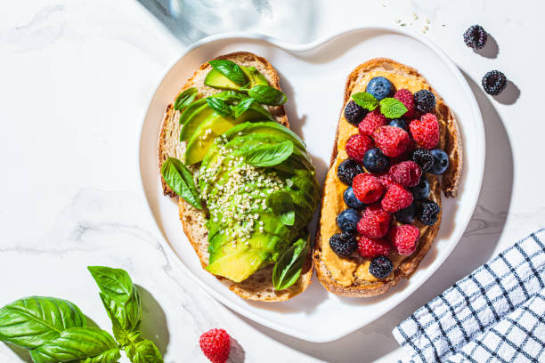 Peanut butter and berries toast and avocado toast with basil on heart plate. Healthy vegan breakfast concept. stock photo