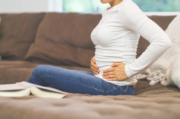 Pelvic pain Young ethnic woman sits on the couch and holds her pelvic area that is in pain pelvis photos stock pictures, royalty-free photos & images